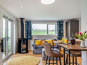 A cozy living room with a fireplace, a sofa with yellow cushions, a dining table with chairs, and a view of a green field through the window. - 1 Bed in Ventnor (oc-91536) (Godshill)