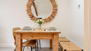 A dining area with a wooden table, chairs, a bench, and a circular mirror. - 1 Bed in Ilfracombe (oc-91652) (Ilfracombe)