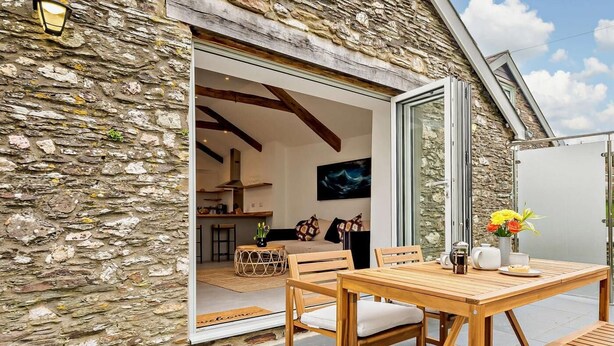 A stone exterior with a wooden dining set and a glass door leading to an interior with a kitchen and seating area. - 1 Bed in Ilfracombe (oc-91652) (Ilfracombe)