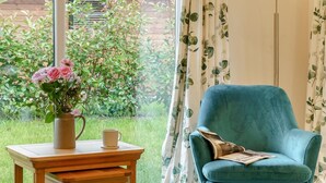 Ein Zimmer mit einem türkisfarbenen Sessel, einem Holztisch mit Blumen und einer Tasse, und Blick auf eine grüne Landschaft.