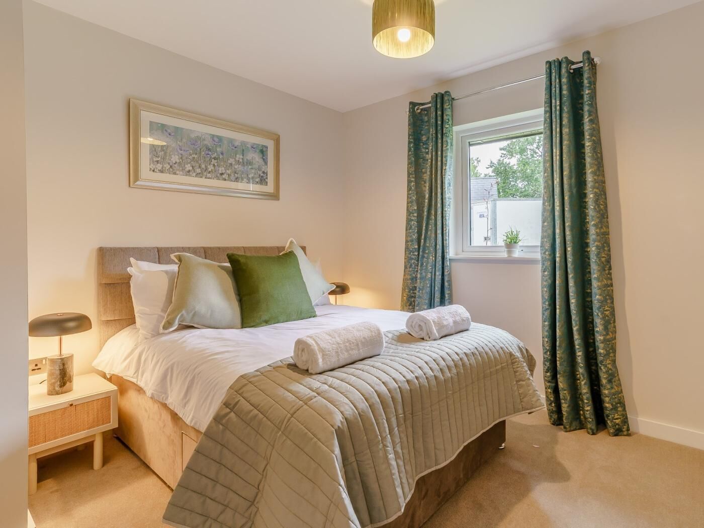 A bedroom with a bed, pillows, a bedside table, a lamp, a window with curtains, and a painting on the wall.