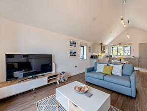 A modern living room with a flat-screen TV, a blue sofa, a coffee table, and a kitchen in the background. - 2 Bed in Bideford (oc-91748) (Northam)