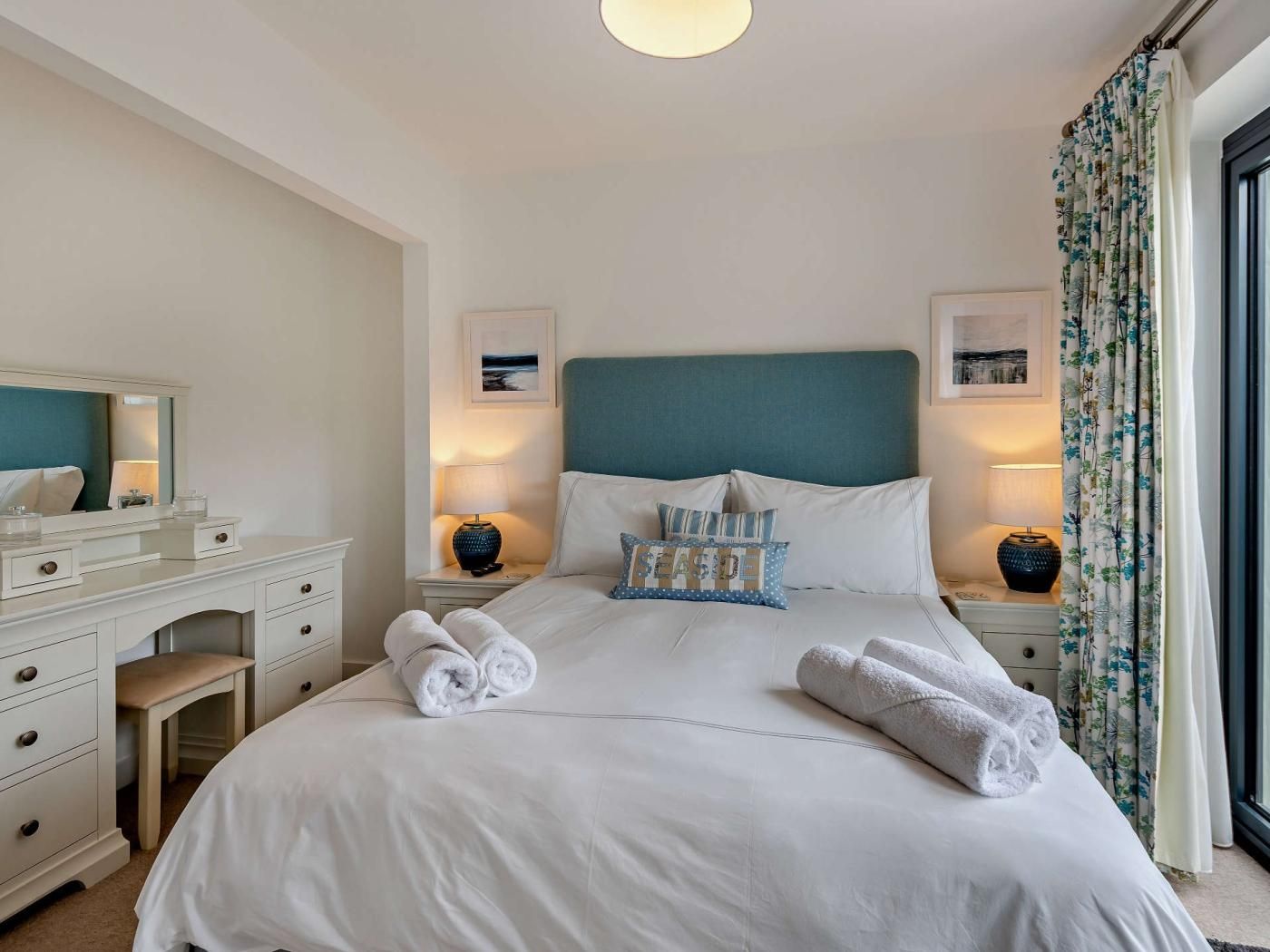 A hotel room with a bed, bedside tables, a dresser, and a window with curtains.