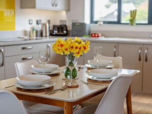 A dining table set with white plates, glasses, and cutlery, a vase with yellow flowers, and a window with a view of greenery. - 2 Bed in Bideford (oc-91748) (Northam)