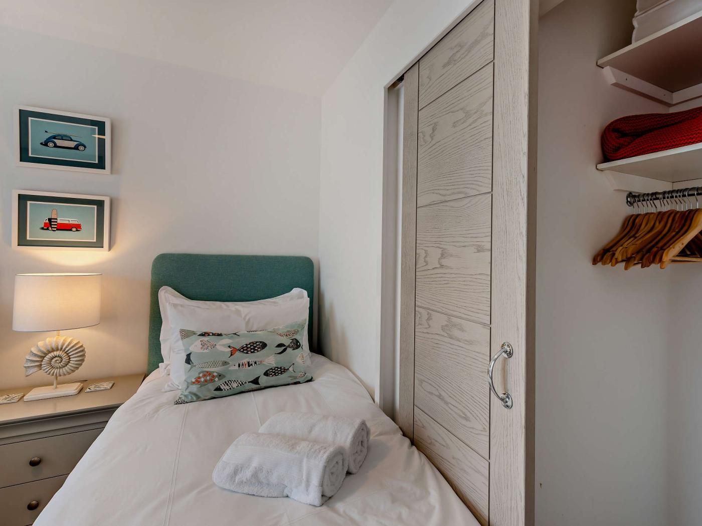 Ein ordentlich bezogenes Bett mit weißen Leinen, ein Türkis-farbener Kopfteil und ein Kissen mit Fischmuster. An der Wand hängen zwei gerahmte Bilder, ein Lampenschirm steht auf dem Nachttisch und ein Holzschrank mit Kleiderstange ist zu sehen.
