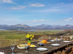 A dining table set outdoors with a scenic view of mountains and a lake. - 6 Bed in Harlech (oc-91546) (Harlech)