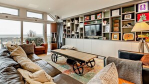 A modern living room with a large sofa, a coffee table, a television, and a bookshelf filled with various items. - 3 Bed in Whitstable (oc-91641) (Seasalter)