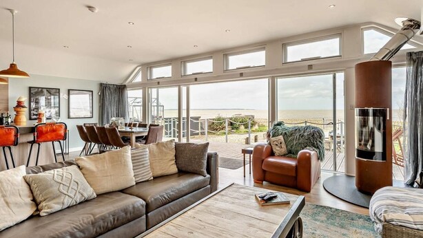 A spacious living area with a large window overlooking the beach, a cozy seating area, and a dining space. - 3 Bed in Whitstable (oc-91641) (Seasalter)