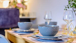 Mesa de jantar posta com pratos, tigelas e talheres, com um vaso de flores ao fundo.