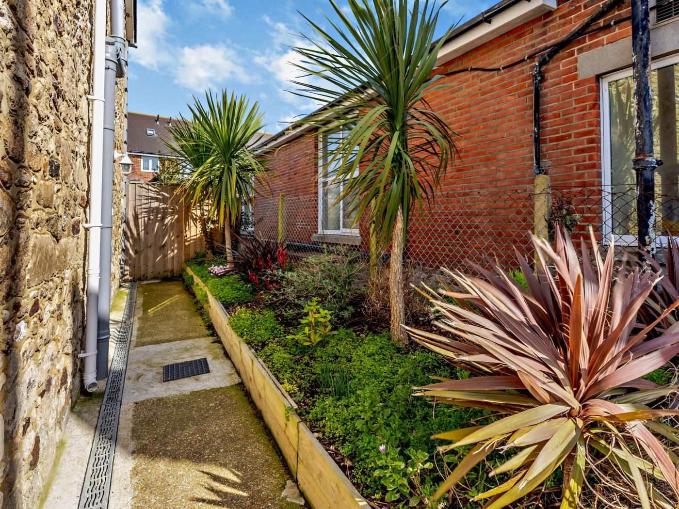 A narrow pathway leading to a brick building with a metal fence and various plants.