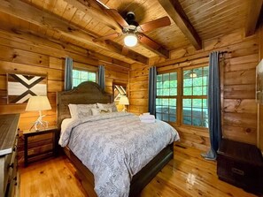 Ein Schlafzimmer in einer Holzhütte mit einem Bett, Nachttischen, einem Deckenventilator und Fenstern mit Vorhängen.