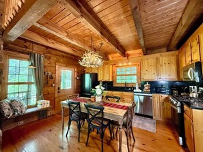A rustic cabin interior with a dining area, wooden ceiling beams, a chandelier, and a kitchen with wooden cabinets. - Boulder Lodge - The Perfect Mountain Escape in Banner Elk near Winery & Hiking (Banner Elk)