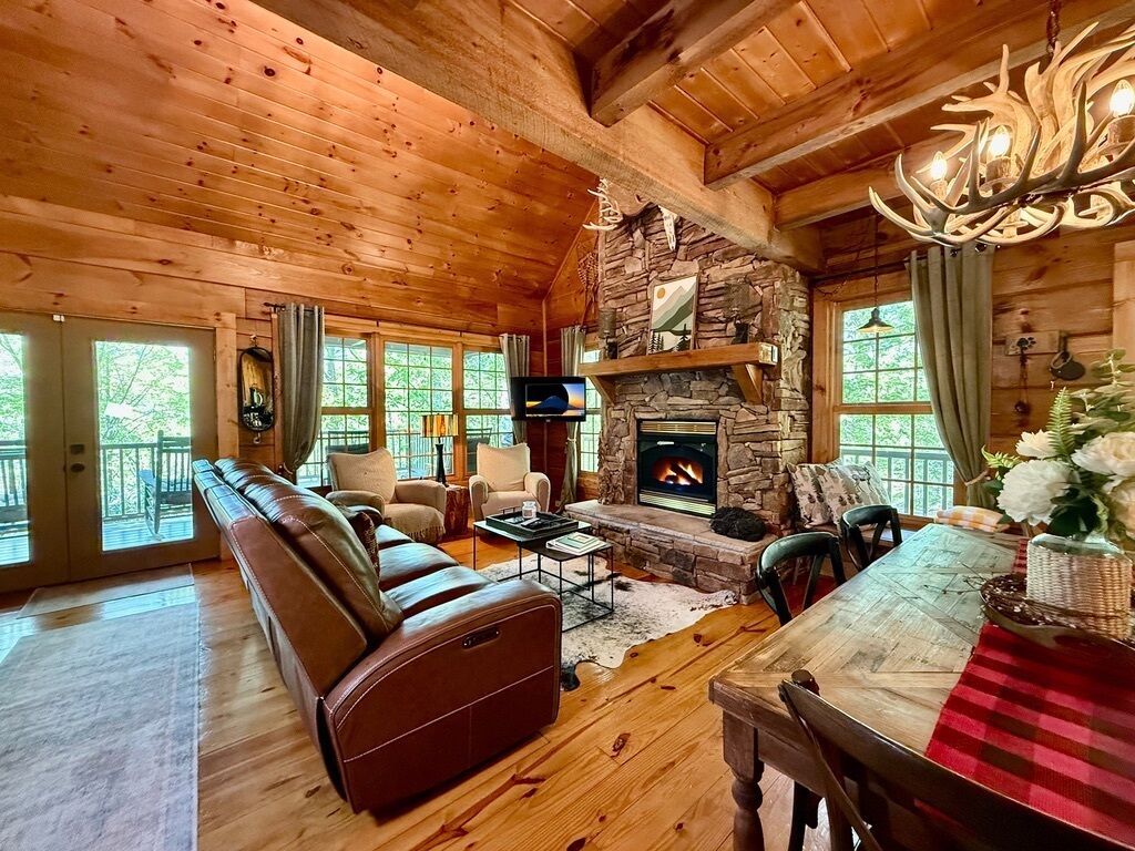 A cozy log cabin interior with a stone fireplace, leather sofa, and a dining area.
