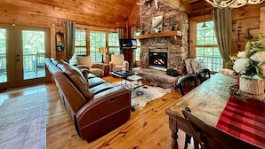 A cozy log cabin interior with a stone fireplace, leather sofa, and a dining area. - Boulder Lodge - The Perfect Mountain Escape in Banner Elk near Winery & Hiking (Banner Elk)
