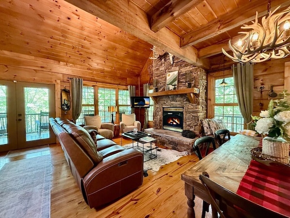 A cozy log cabin interior with a stone fireplace, leather sofa, and a dining area. - Boulder Lodge - The Perfect Mountain Escape in Banner Elk near Winery & Hiking (Banner Elk)