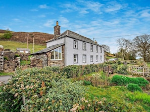 A two-story house with a stone chimney, a large window, and a garden with various plants. - 1 Bed in Penrith (oc-91812) (Ullswater)