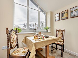 A dining area with a table set for two, chairs with floral upholstery, a window with a view of trees, and framed pictures on the wall. - 1 Bed in Penrith (oc-91812) (Ullswater)