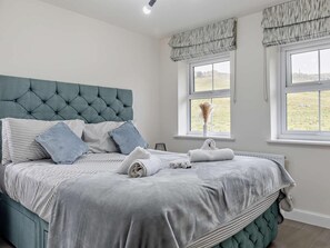 A bedroom with a tufted headboard, striped bedding, and two windows with curtains. - 3 Bed in Buxton (oc-91785) (Buxton)