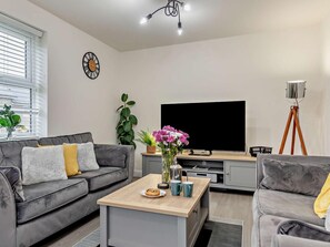 A modern living room with a grey sofa, a coffee table with a plate of food, a TV, and a plant. - 3 Bed in Buxton (oc-91785) (Buxton)