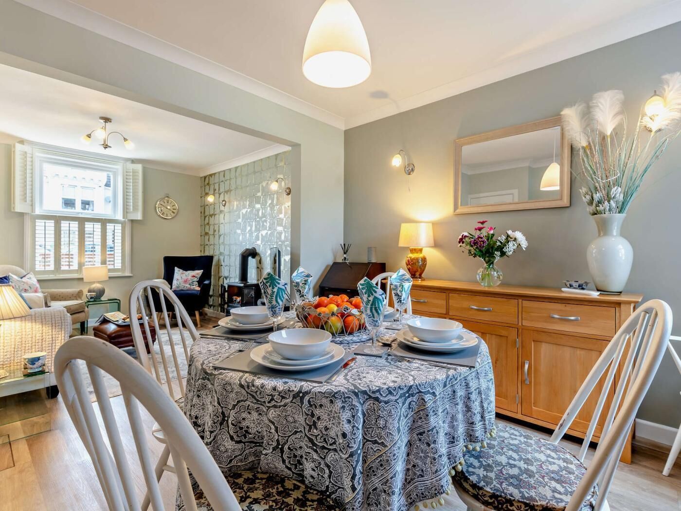 A dining area with a patterned tablecloth, white chairs, a wooden cabinet, and a mirror on the wall.