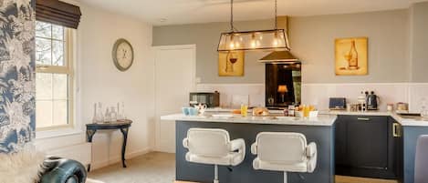 A modern kitchen with a central island, two white bar stools, a clock on the wall, and a floral arrangement on the floor.