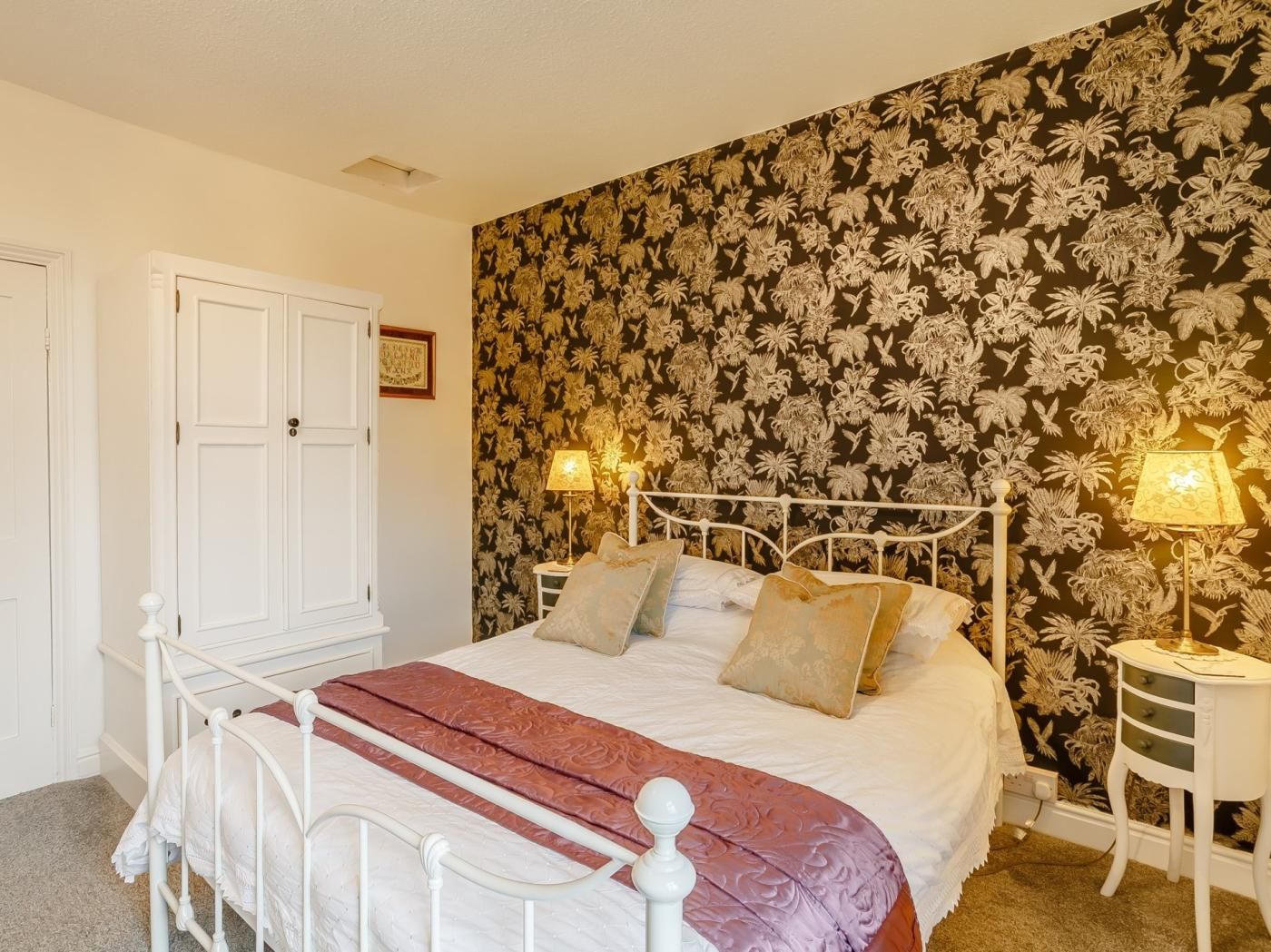 A bedroom with a white metal bed frame, a floral wallpaper, two bedside lamps, and a white bedside table.
