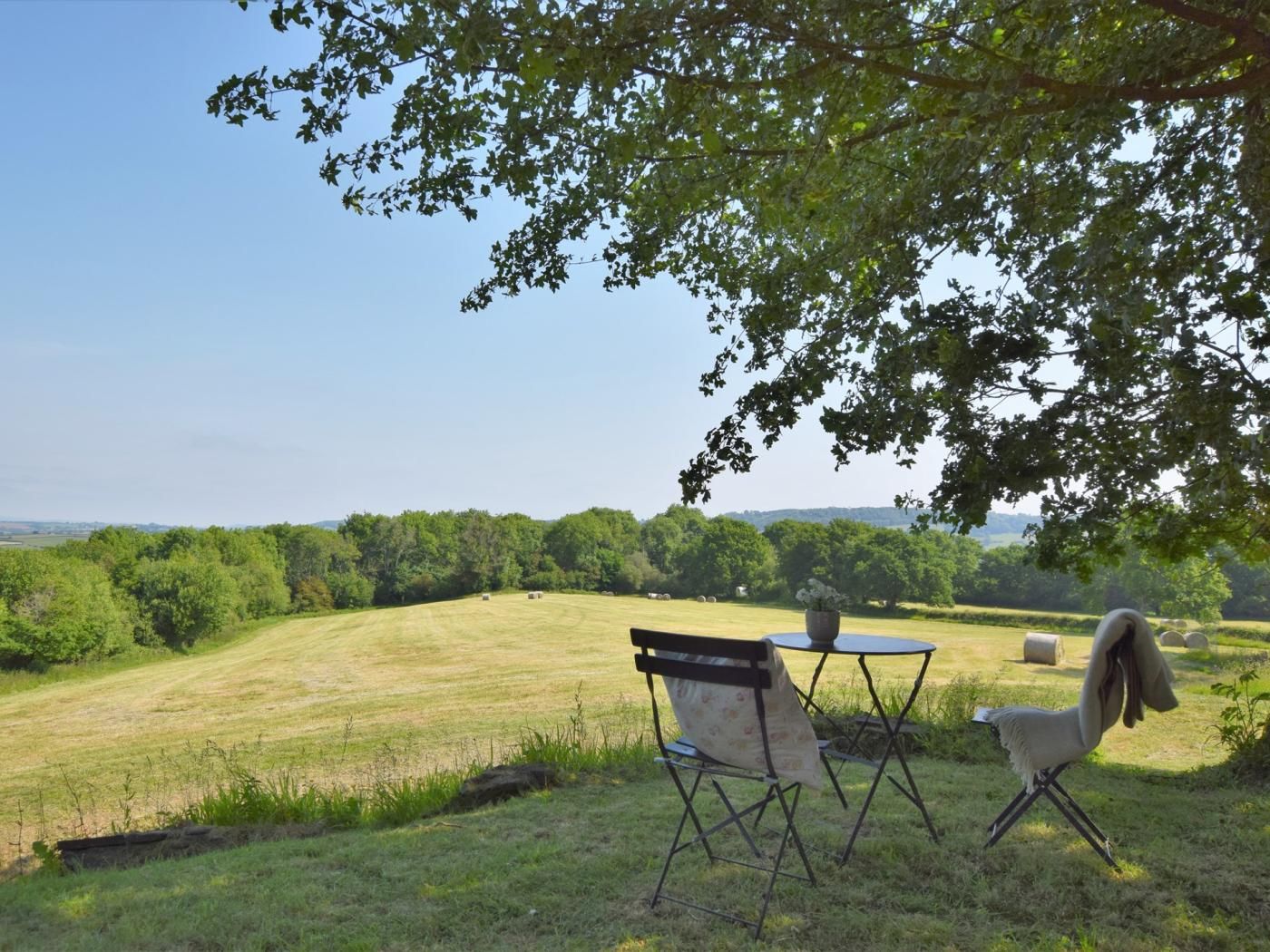 An outdoor seating area with a table and chairs under a tree, overlooking a grassy field with hay bales.