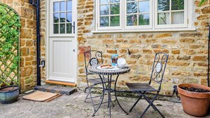 A stone building with a white door and windows, a small round table with chairs, and a potted plant. - 2 Bed in Banbury (oc-92008) (Hook Norton)