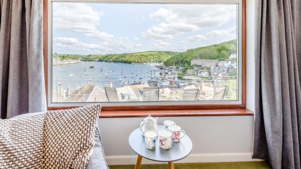 A room with a large window overlooking a harbor, a bed with patterned pillows, and a small table with teacups. - 2 Bed in Fowey (oc-92080) (Fowey)