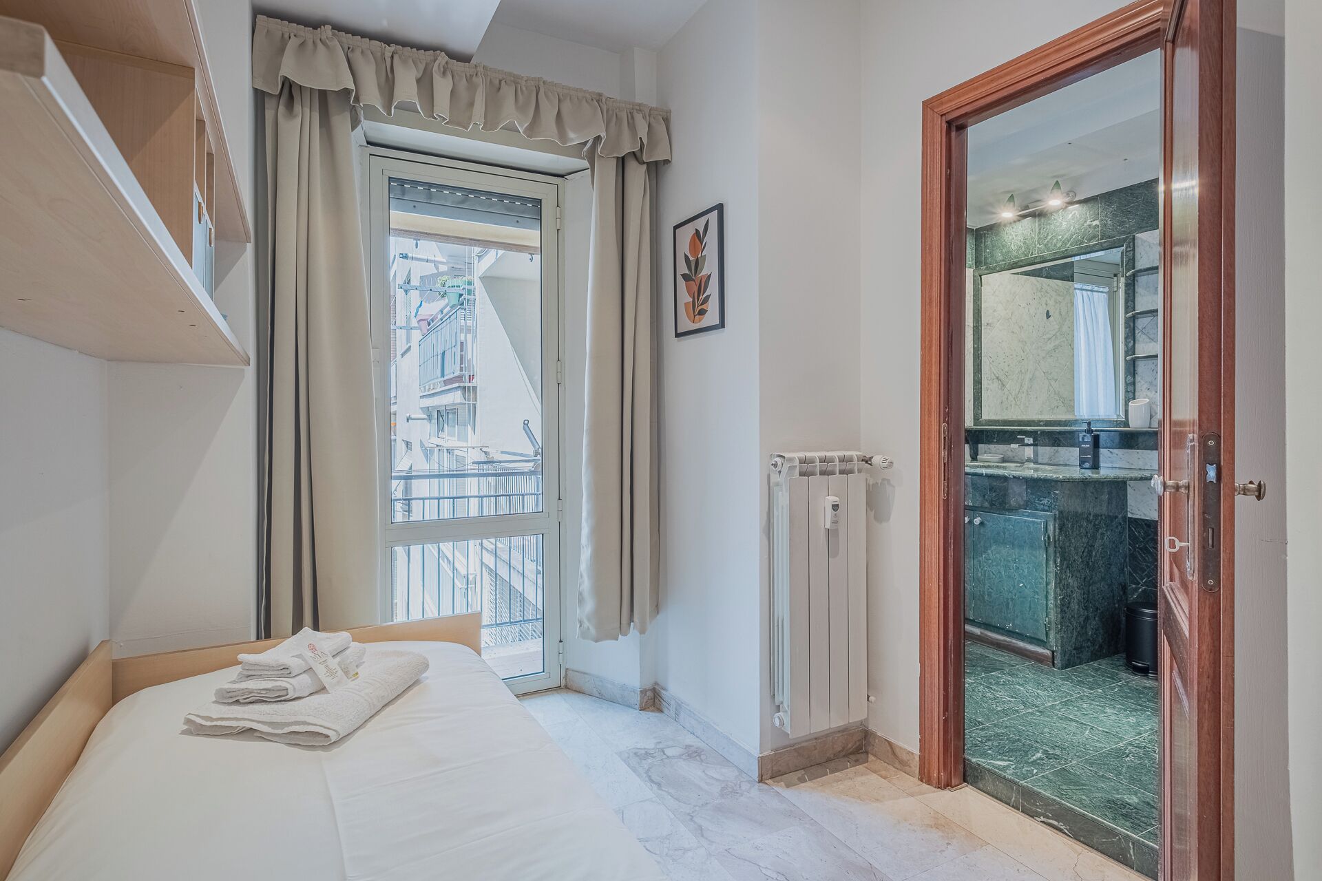 Un dormitorio con cama, ventana con cortinas, radiador y baño visible a través de una puerta abierta.