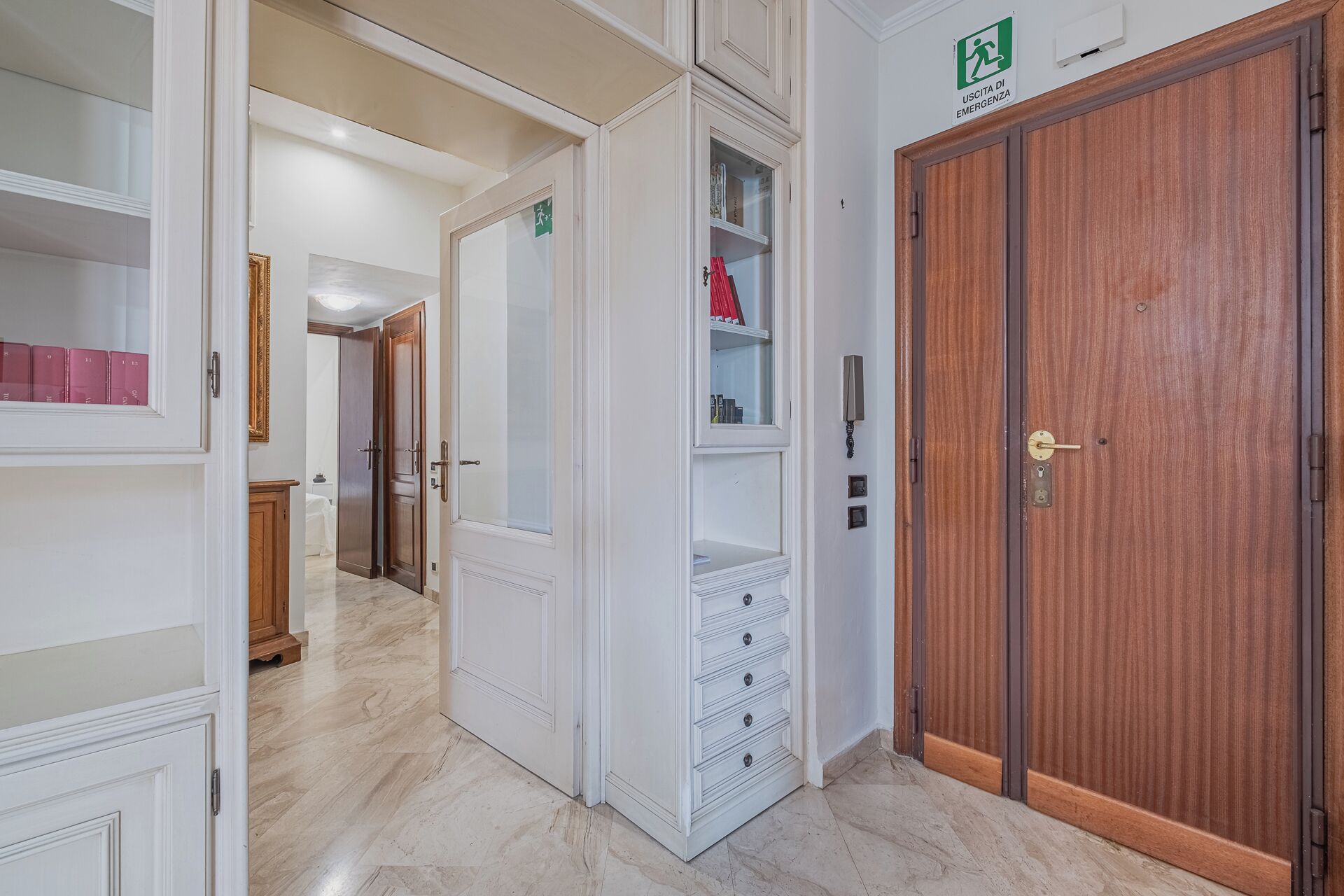 A hotel corridor with a wooden door, a white cabinet with drawers, and a glass-fronted shelf unit.