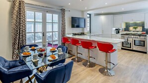 Une cuisine moderne avec des meubles blancs, un îlot central doté de tabourets de bar et un coin repas avec une table en verre et des chaises bleues.