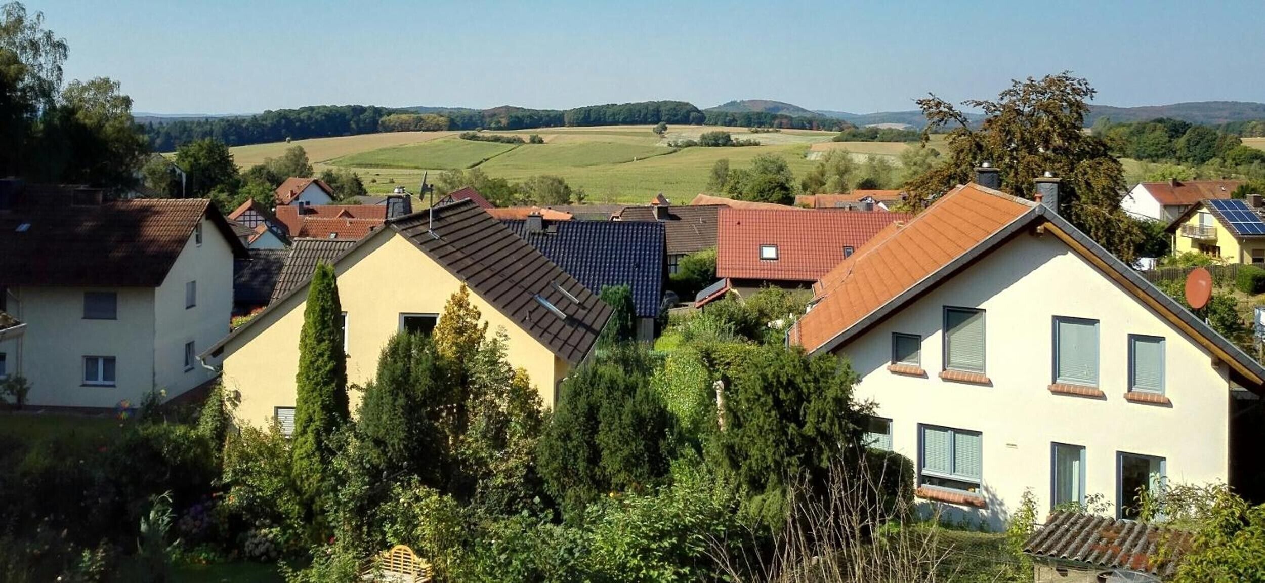 Zona residencial con casas, algunas con paneles solares, rodeada de vegetación y colinas al fondo.