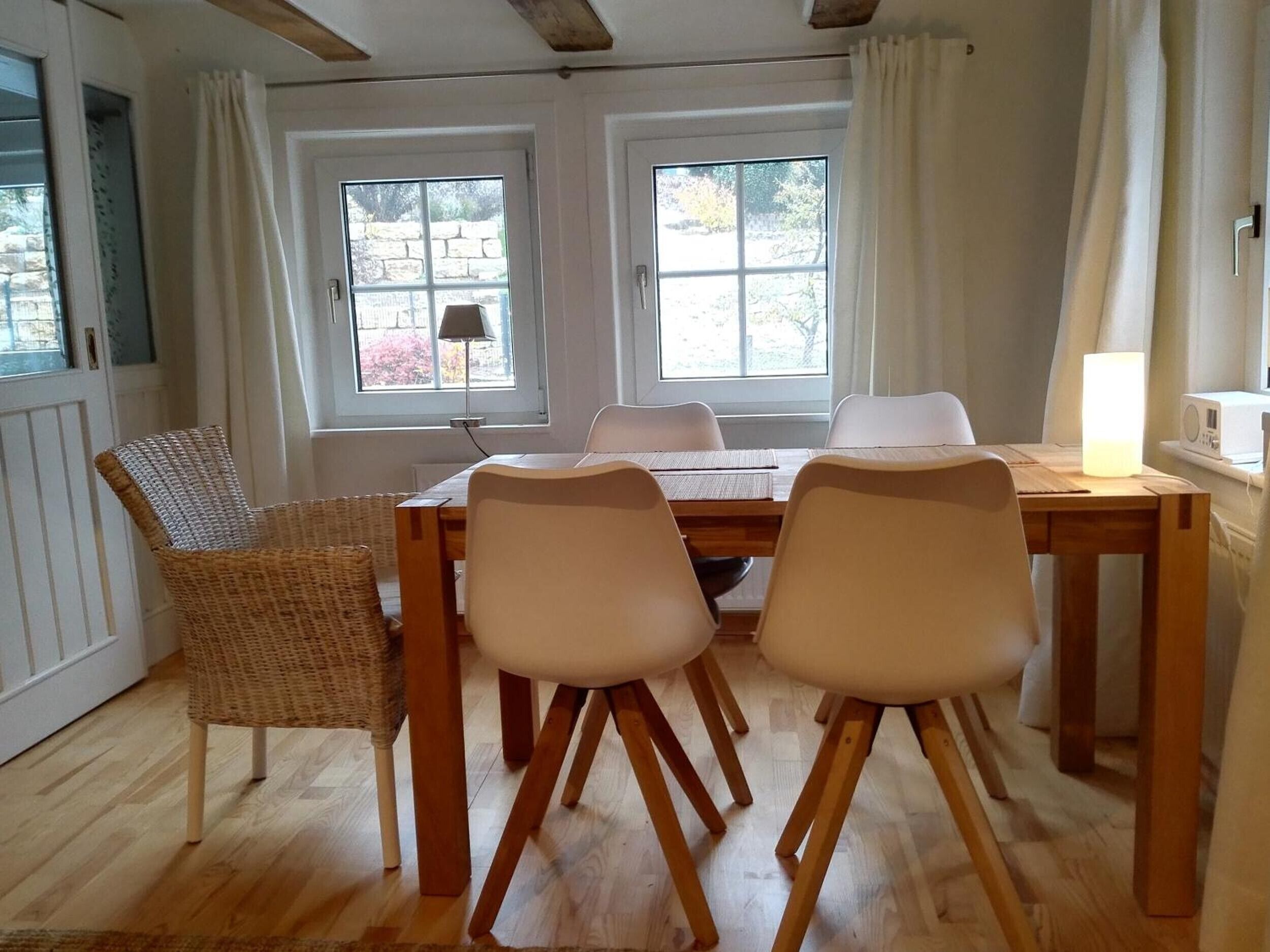 A dining area with a wooden table and four chairs, two windows with curtains, and a wicker chair.