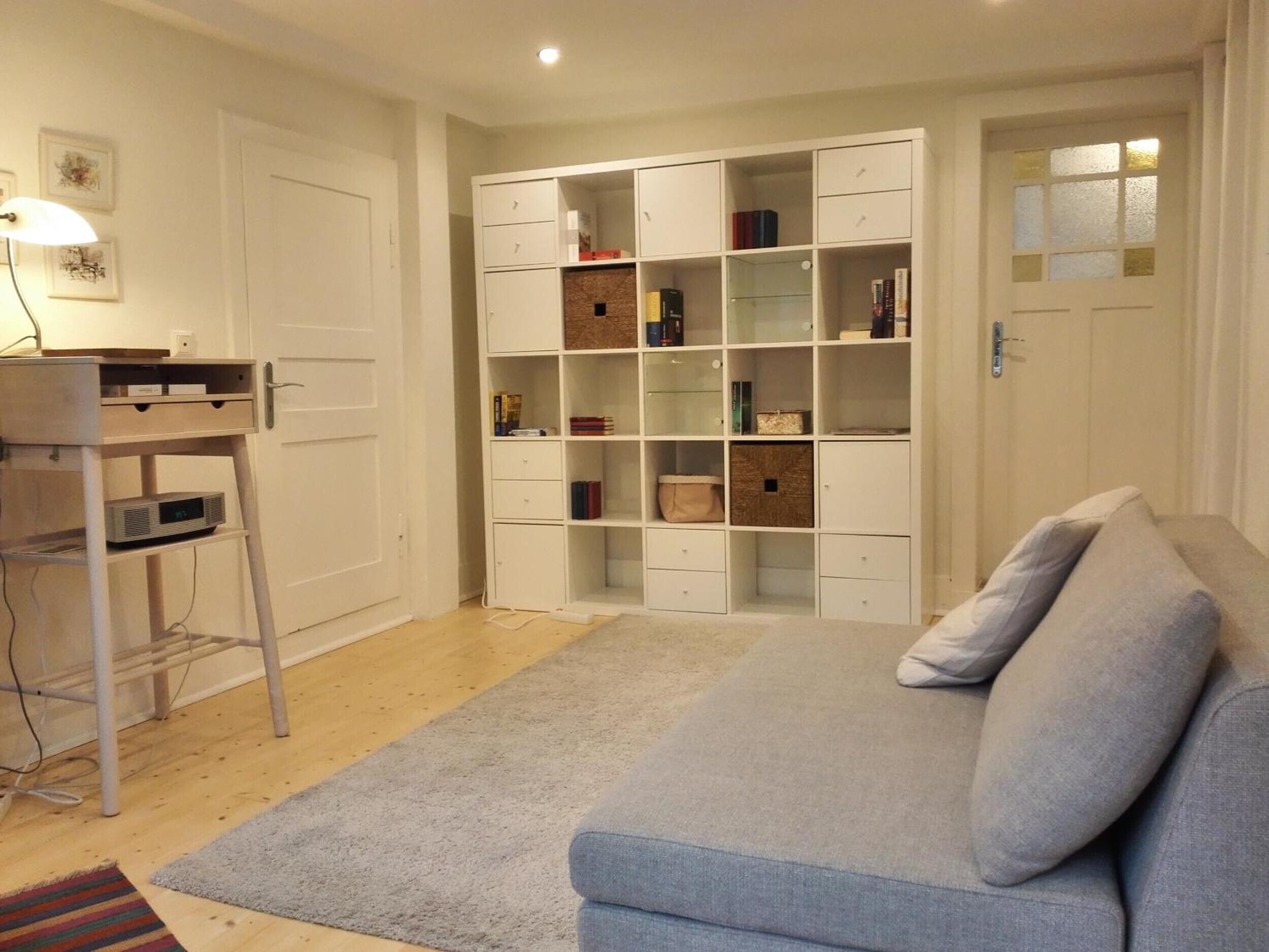 Une pièce avec un canapé gris, un meuble de rangement blanc, un bureau en bois avec une lampe et un tapis.