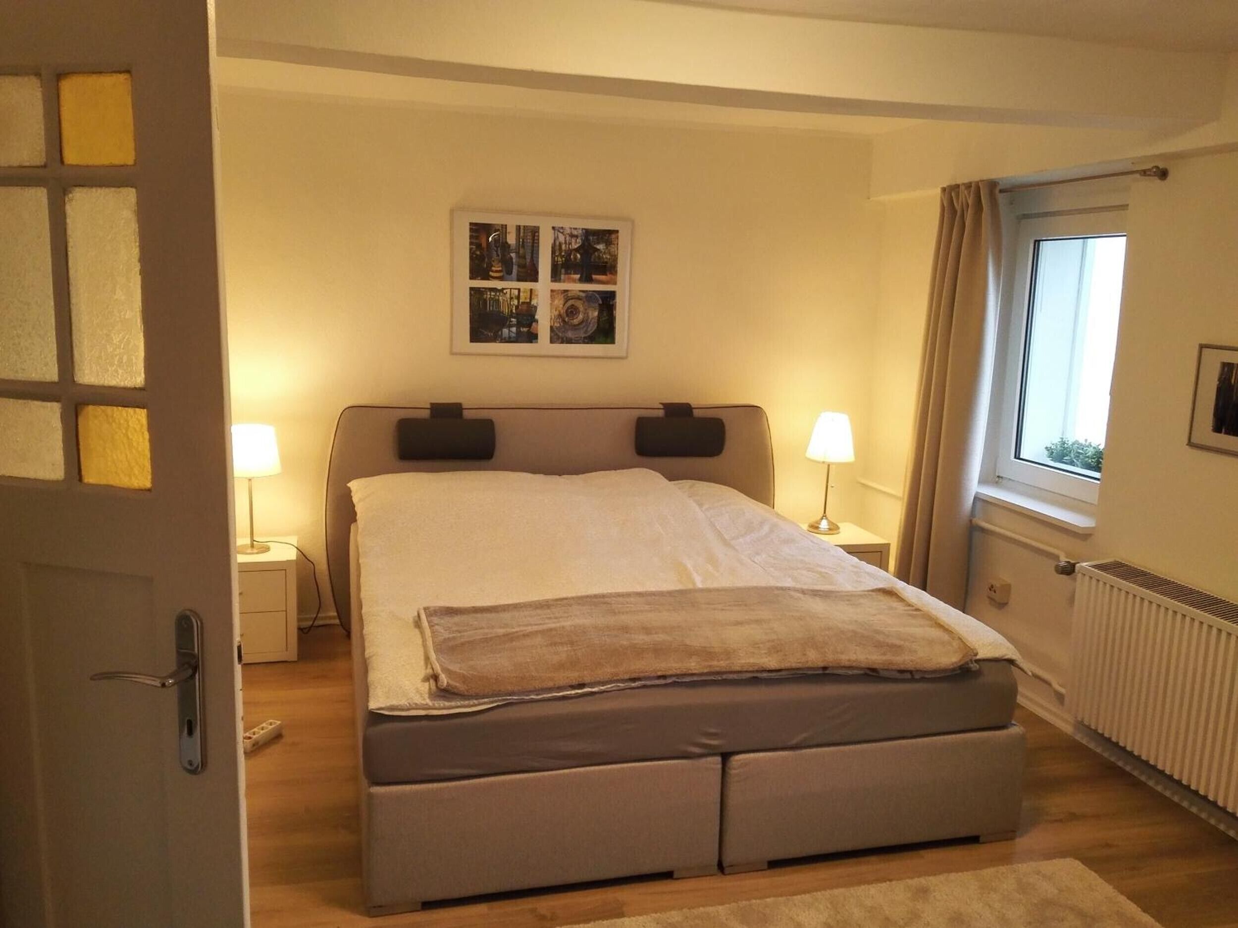 Une chambre à coucher avec un lit, des lampes de chevet, une fenêtre avec des rideaux, un radiateur et une porte.