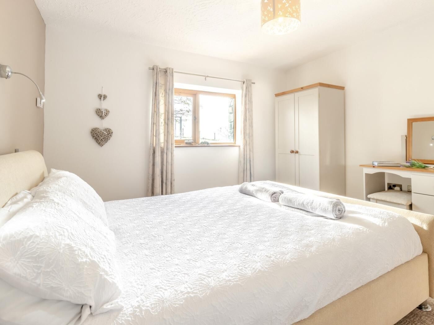 Un dormitorio con cama, mesita de noche, armario, ventana con cortinas y lámpara de pared.