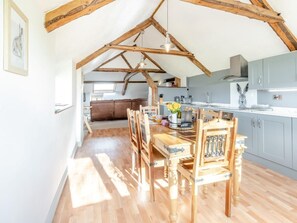 Ein Essbereich mit Holztisch und Stühlen, eine Küche im Hintergrund und eine loftartige Decke mit freiliegenden Balken.