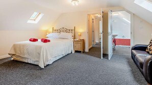 Ein Schlafzimmer mit einem Bett, einem Nachttisch, einer Nachttischlampe, einem roten Handtuch, einem schwarzen Ledersessel und einem durchgehenden Türspalt einsehbaren Badezimmer.