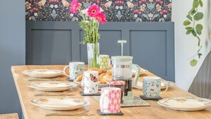 Una tavola imbandita per la colazione con piatti, una caffettiera, un vaso con fiori e una decorazione a parete.