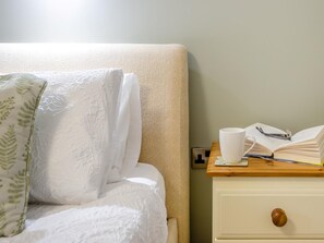 Ein ordentlich bezogenes Bett mit einem weißen Kissen und einem Kissen mit grünem Blattmuster, ein Nachttisch mit Buch, Brille und Tasse sowie ein kleiner Holzbeistelltisch mit Schublade.