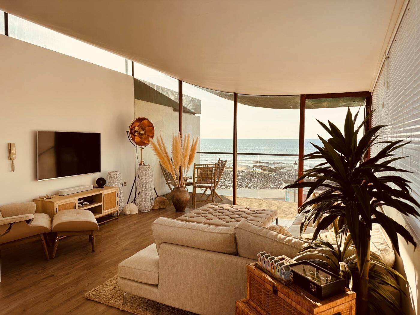 A modern living room with a large TV, comfortable seating, a fan, and a view of the beach.