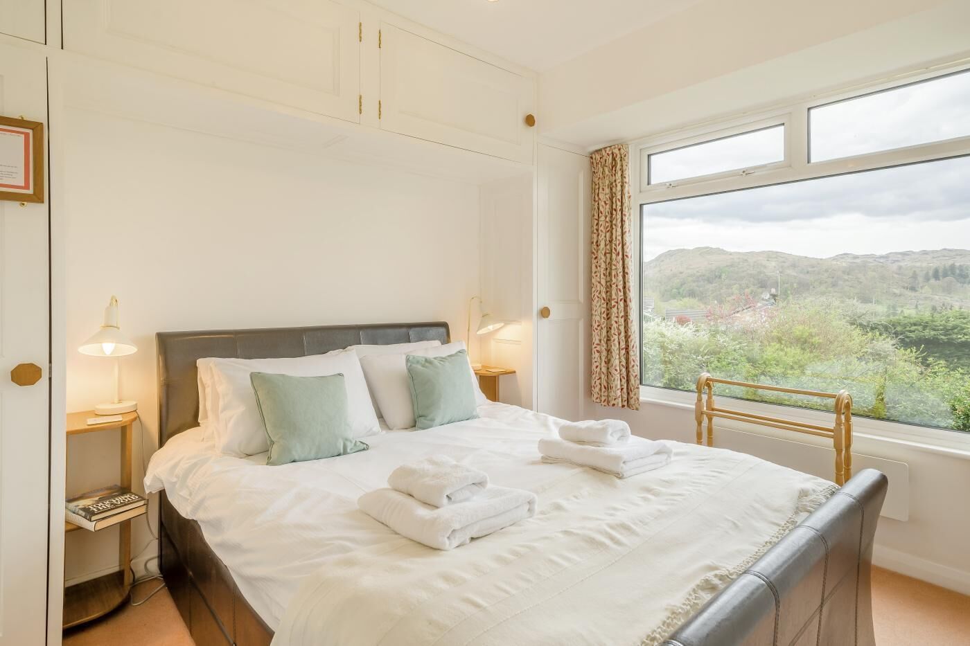 A neatly made bed with pillows, a bedside table with a lamp, and a window with a view of greenery.