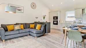 Ein modernes Wohnzimmer mit einem grauen Ecksofa, einem Essbereich mit Holztisch und Stühlen, einer Küche mit weißen Schränken und Geräten sowie einer dekorativen Wanduhr.
