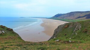 A coastal landscape with a beach, grassy hills, and a distant view of the sea. - 3 Bed in Swansea (oc-91721) (Upper Killay)