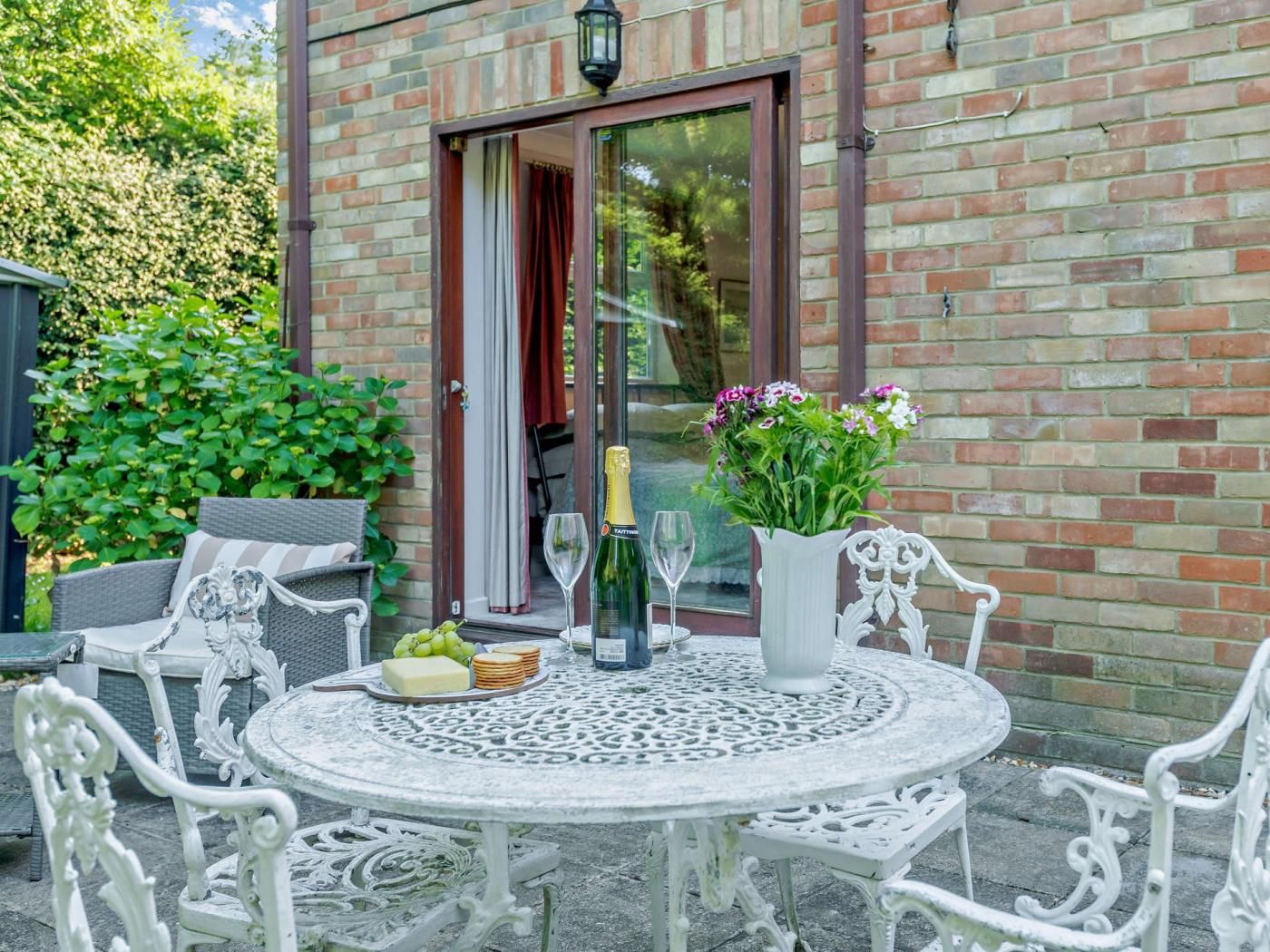 An outdoor patio with a round table, white chairs, a bottle of champagne, a vase with flowers, and a glass.