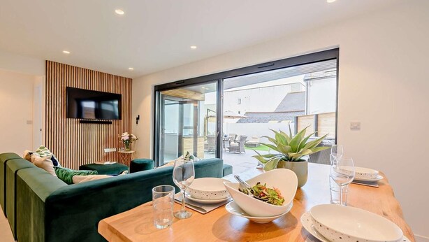 Una sala de estar moderna con un sofá, una mesa de comedor con platos y vasos, un televisor en la pared y vistas al exterior.