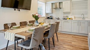 Un coin repas avec un téléviseur fixé au mur, une cuisine avec des meubles blancs et une table à manger dressée avec des assiettes et des verres.