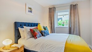 Une chambre à coucher avec un lit, une table de chevet avec une lampe, une fenêtre avec des rideaux et un tableau accroché au mur.
