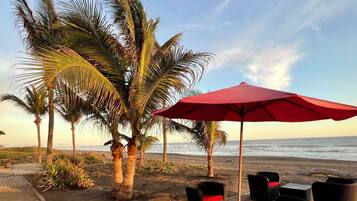Une terrasse au bord de la plage, avec des meubles en osier et un parasol rouge, des palmiers et une vue sur l’océan.